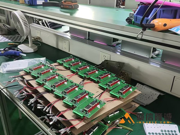 电子电路板在生产线上排列整齐，连接着红色导线，背景为工厂工作台和工具。